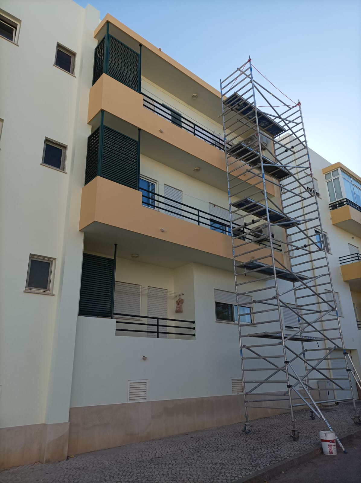 Luxury Villa Renovation Algarve. A low-angle view of a multi-story residential building undergoing exterior renovation. The building has white and light peach-colored facades and black railings on the balconies. A tall, modular metal scaffolding structure is erected against the side of the building, reaching several floors, indicating construction or maintenance work.