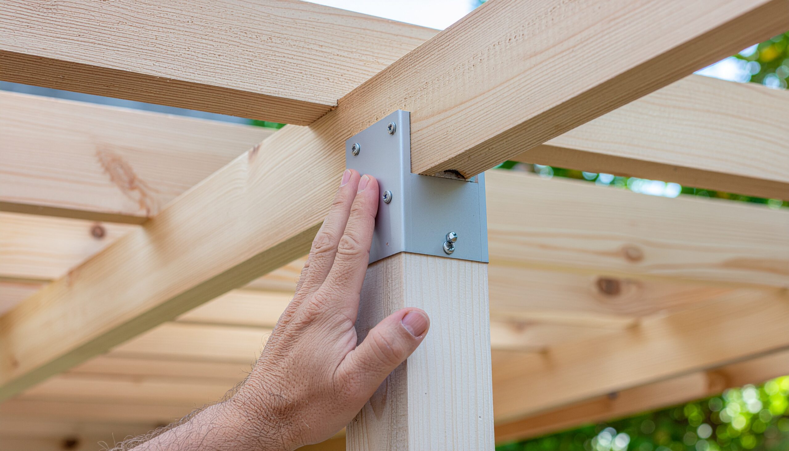 Close-up shot of a person's arm and hand holding a metal joining plate at the light wood corner of a pergola under construction. The plate is secured with screws, connecting a horizontal beam to a vertical post. A pale sky and white roof sections are visible in the background.