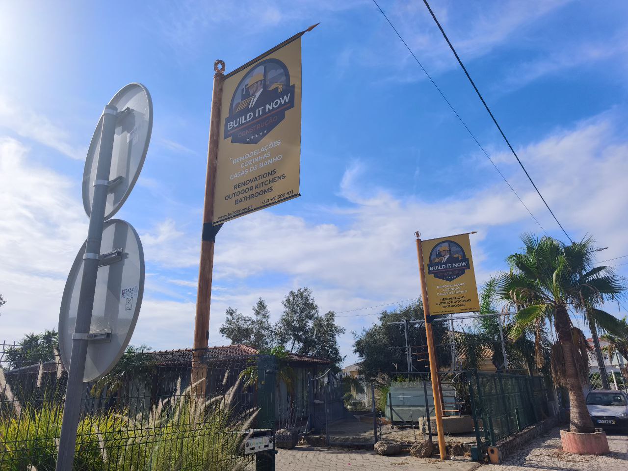 Low-angle shot of a two-lane rural road with a cobblestone pavement and sidewalk on the left, under a blue sky with white clouds. Two large yellow advertising banners for the company "Build It Now" are visible on the left side of the road. Some buildings, vegetation, and a couple of cars are seen in the distance.