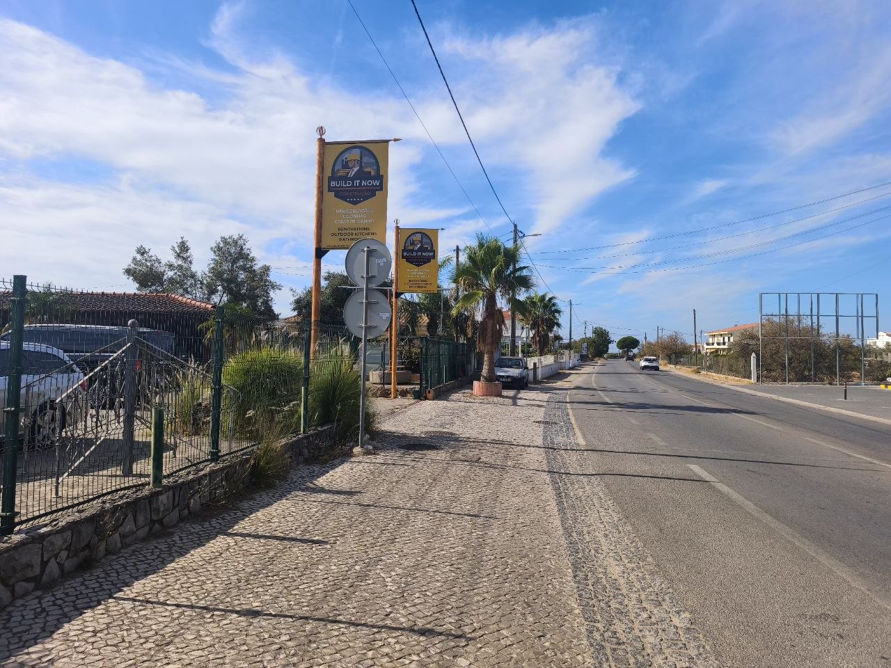 Close-up of two yellow advertising banners for the company "Build It Now" featuring text in Portuguese and English (Remodelações, Cozinhas, Casas de Banho / Renovations, Outdoor Kitchens, Bathrooms). The banners are mounted on wooden poles. To the left, two round traffic signs are visible. The background shows vegetation, a low wall, and a bright blue sky with clouds.