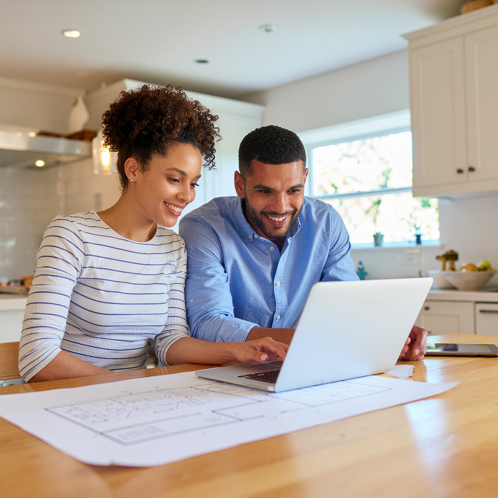 A smiling couple reviewing blueprints and architectural drawings on a laptop in their bright kitchen, symbolizing project consultation, design, and collaboration.