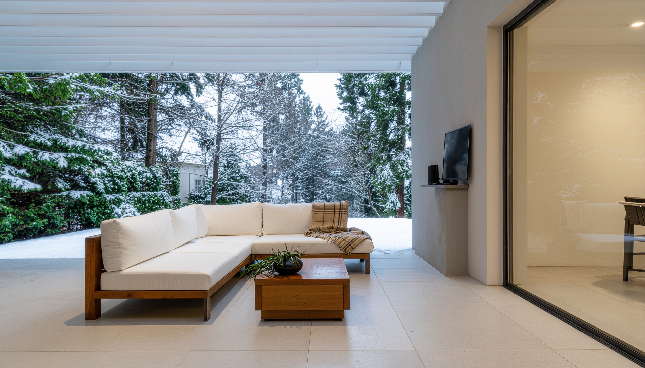 Modern outdoor living room featuring white wooden sectional sofas, a wood coffee table, and a throw blanket, situated on a covered patio that provides shelter from a snowy landscape of trees in the background.