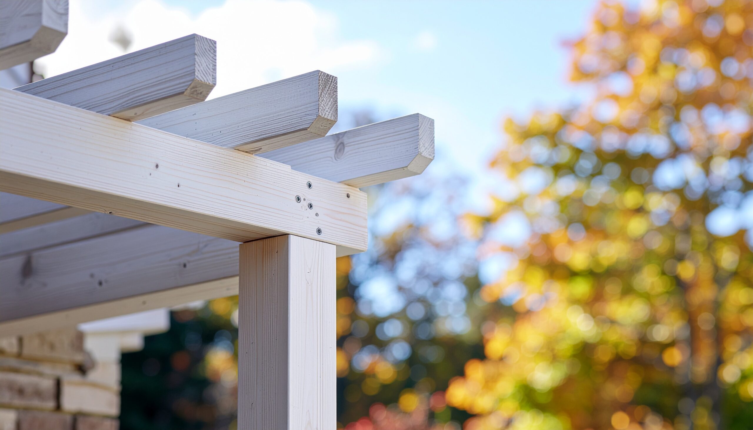 Close-up detail of a white painted wooden pergola beam and rafter joint, showcasing the construction quality and finish, set against a blurred background of yellow and orange autumn foliage under a blue sky.