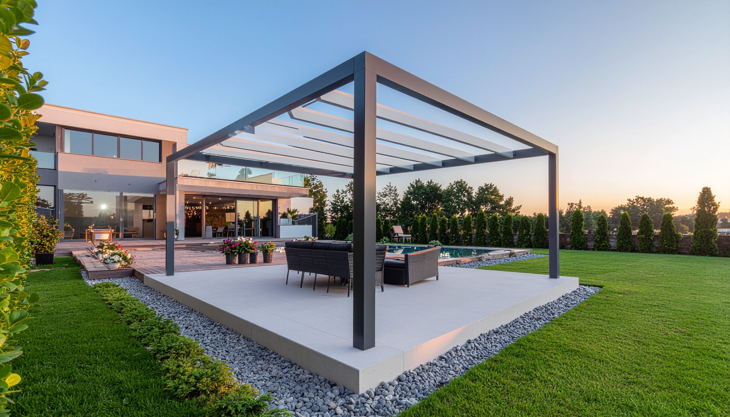 Minimalist black aluminum pergola with a retractable louvered roof, set on a raised concrete patio in a lush green lawn, next to a contemporary luxury home at sunset.
