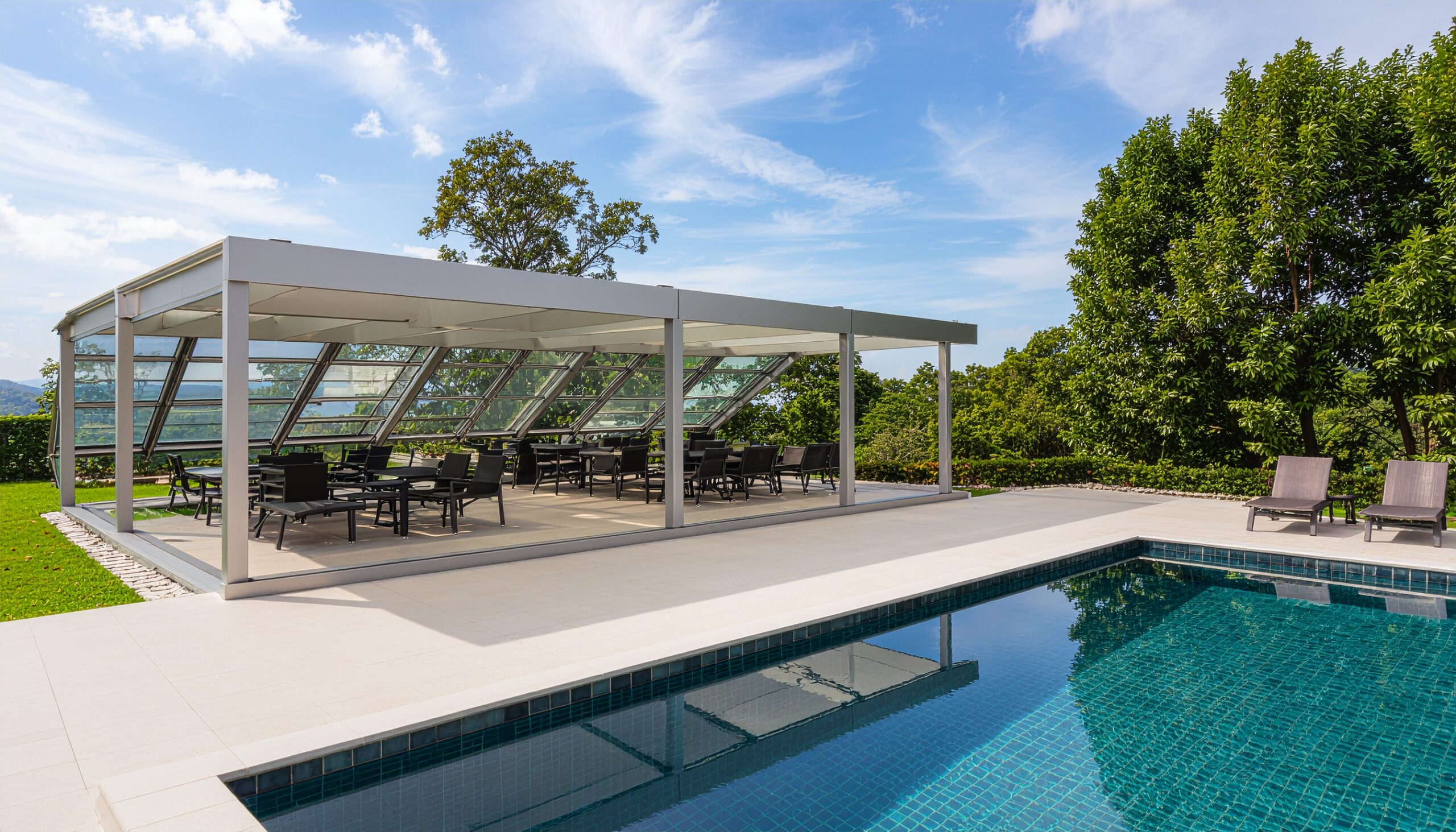 Large aluminum pergola with clear glass sliding walls fully opened, providing an enclosed dining or event space next to a rectangular swimming pool with dark tile, under a sunny, blue sky.