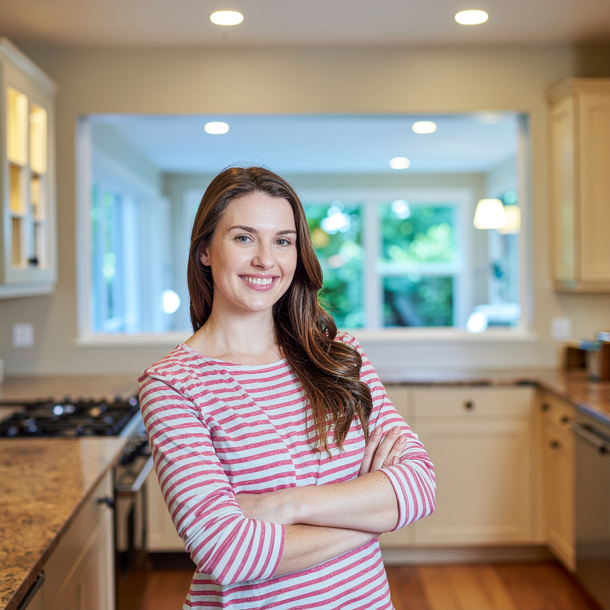 Portrait of a smiling Caucasian woman with her arms crossed, standing in a bright, modern kitchen with granite countertops and a window overlooking a garden, suitable for representing customer satisfaction or a project consultant.