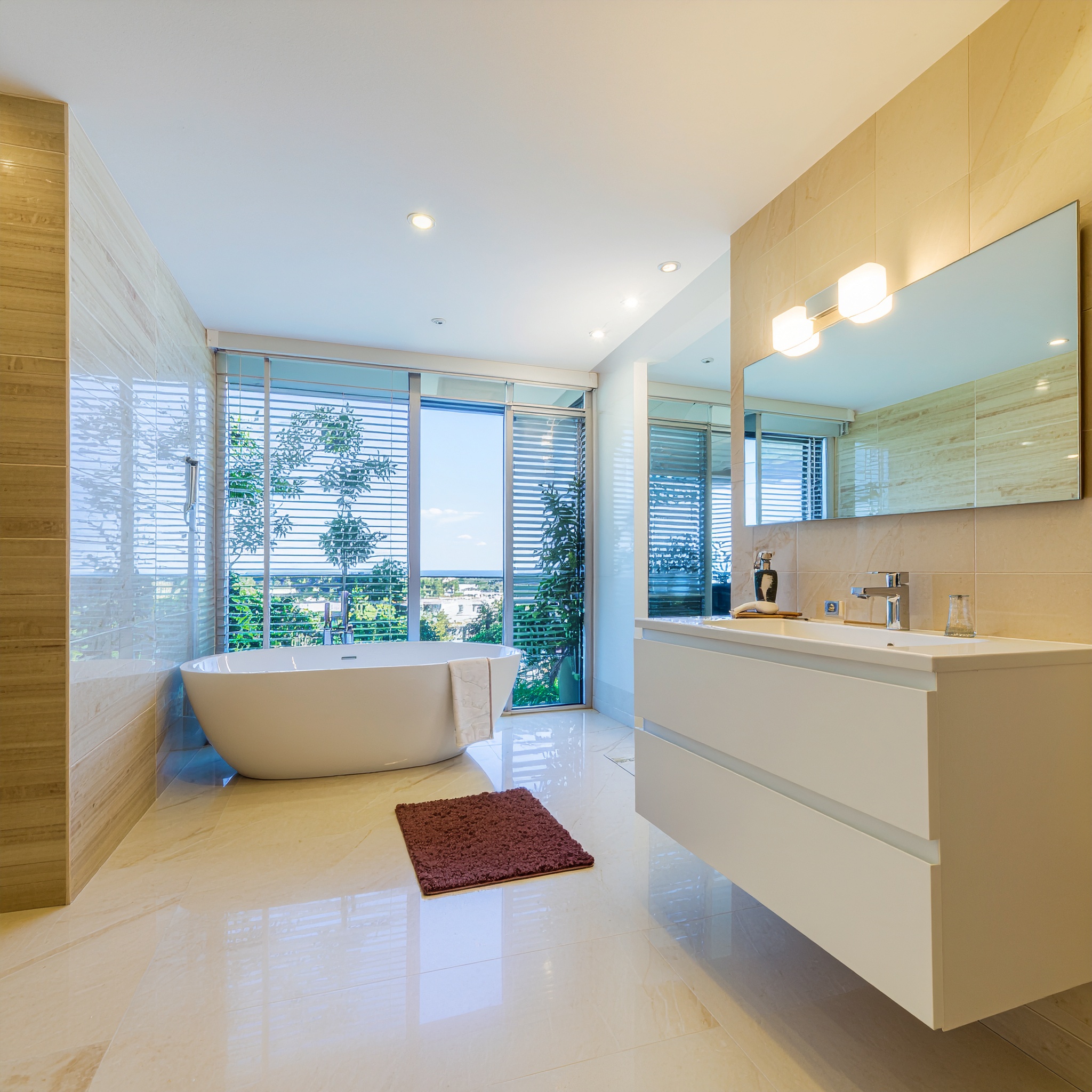 A bright, modern luxury bathroom featuring polished cream-colored floor and wall tiles. A white floating vanity with a single sink and a large rectangular mirror dominates the right side. On the left, a white freestanding bathtub sits in front of a floor-to-ceiling window covered by horizontal blinds, offering a view of greenery. A small, dark red bath mat is placed beside the tub.