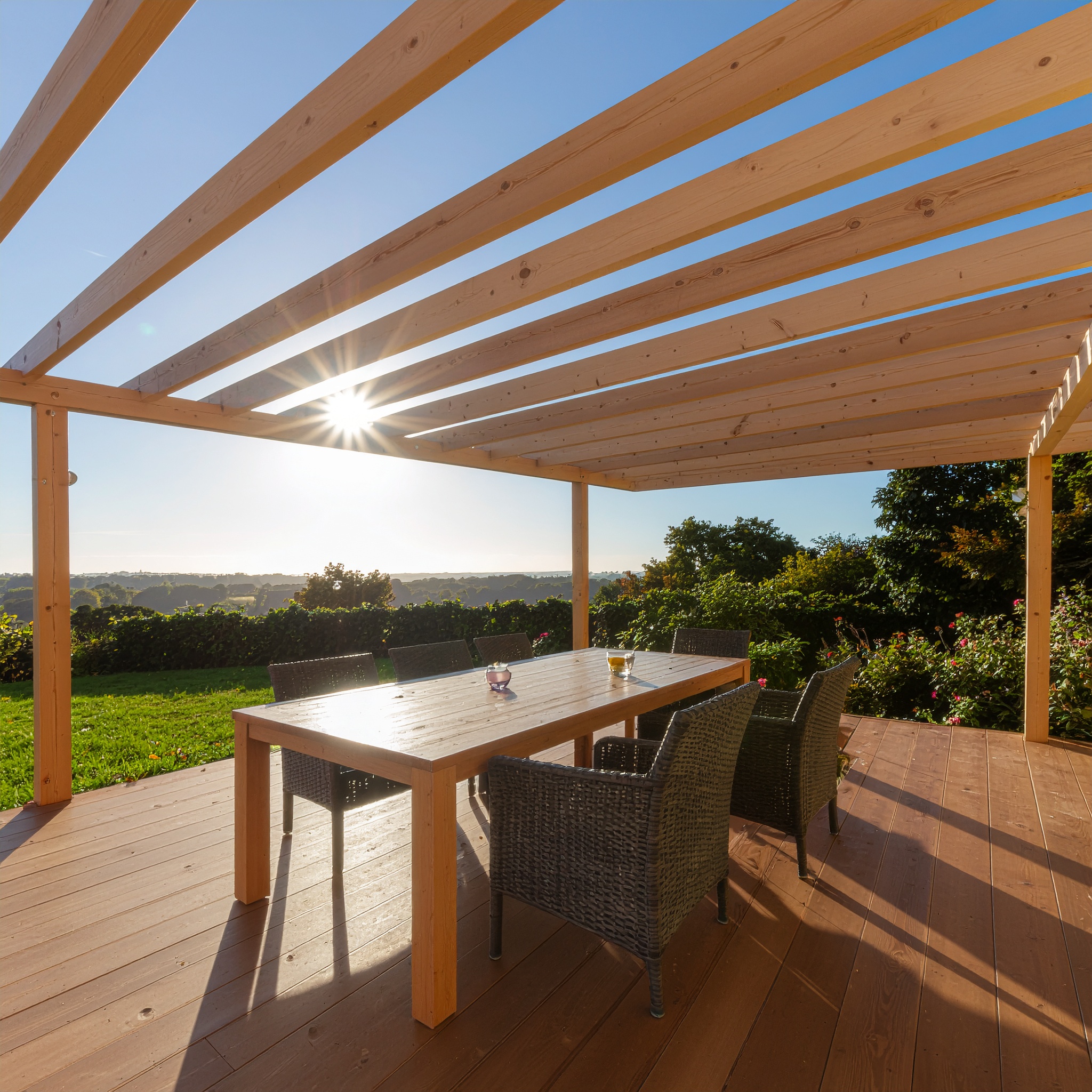 A newly constructed, unfinished natural wooden pergola with spaced rafters, covering a modern outdoor dining table and wicker chairs on a wooden deck, with bright sunlight streaming through the structure, overlooking a lush green lawn.