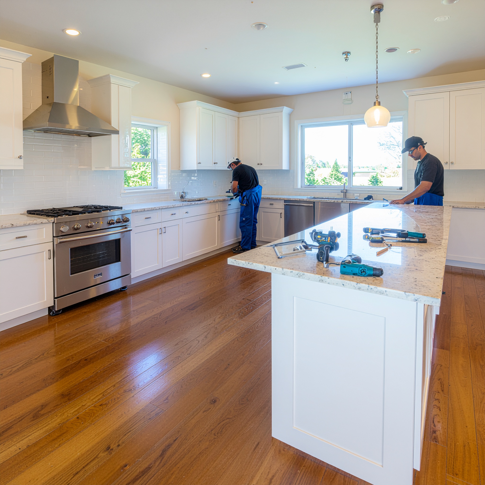 Two workers in uniform installing appliances and cabinetry in a newly renovated, bright white kitchen with granite countertops, stainless steel appliances, and polished hardwood floors, demonstrating professional installation services.