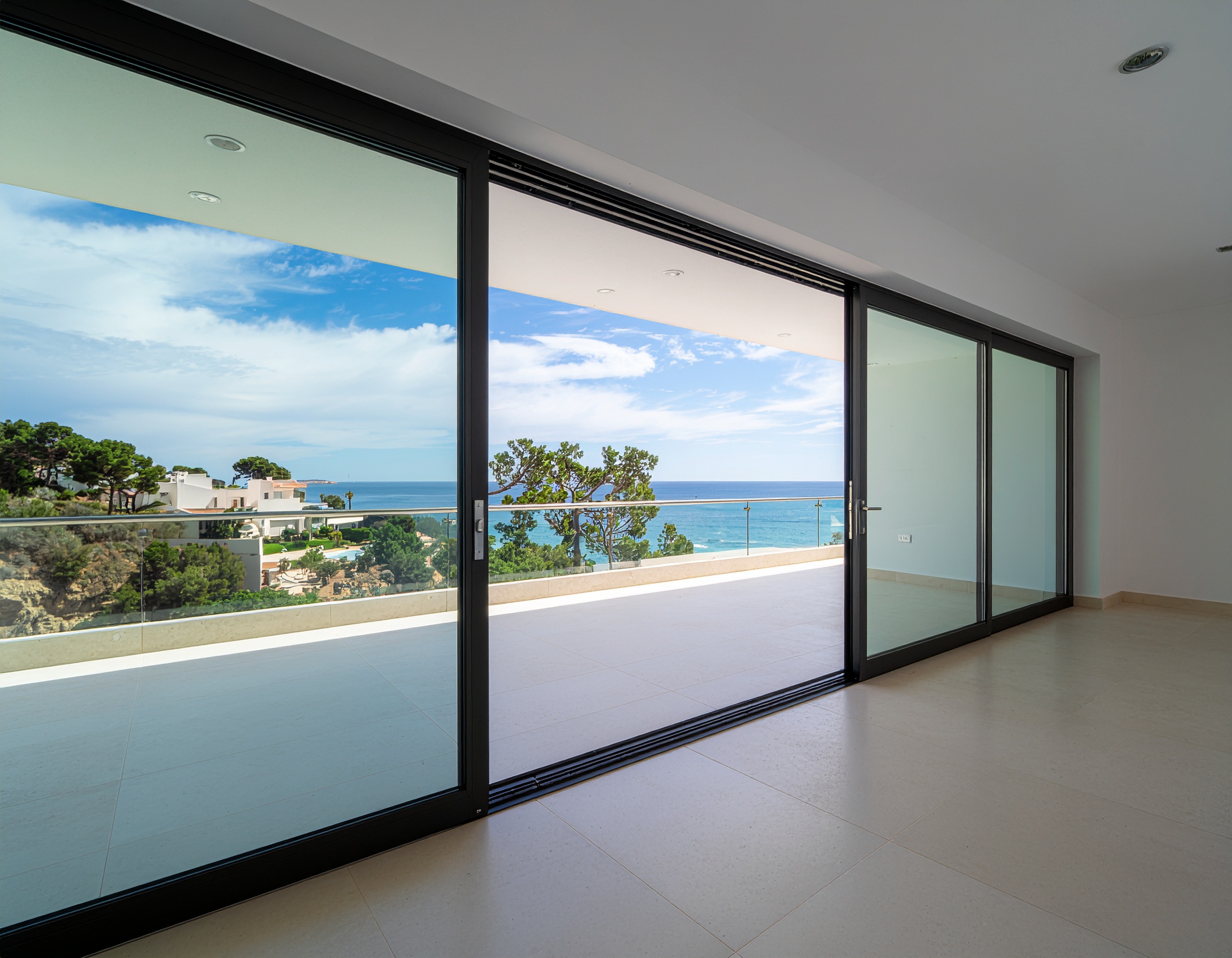 Interior view of a room with large, multi-panel black-framed sliding glass doors opening onto an expansive outdoor terrace with a glass railing. The view overlooks the ocean, green trees, and distant white coastal homes.