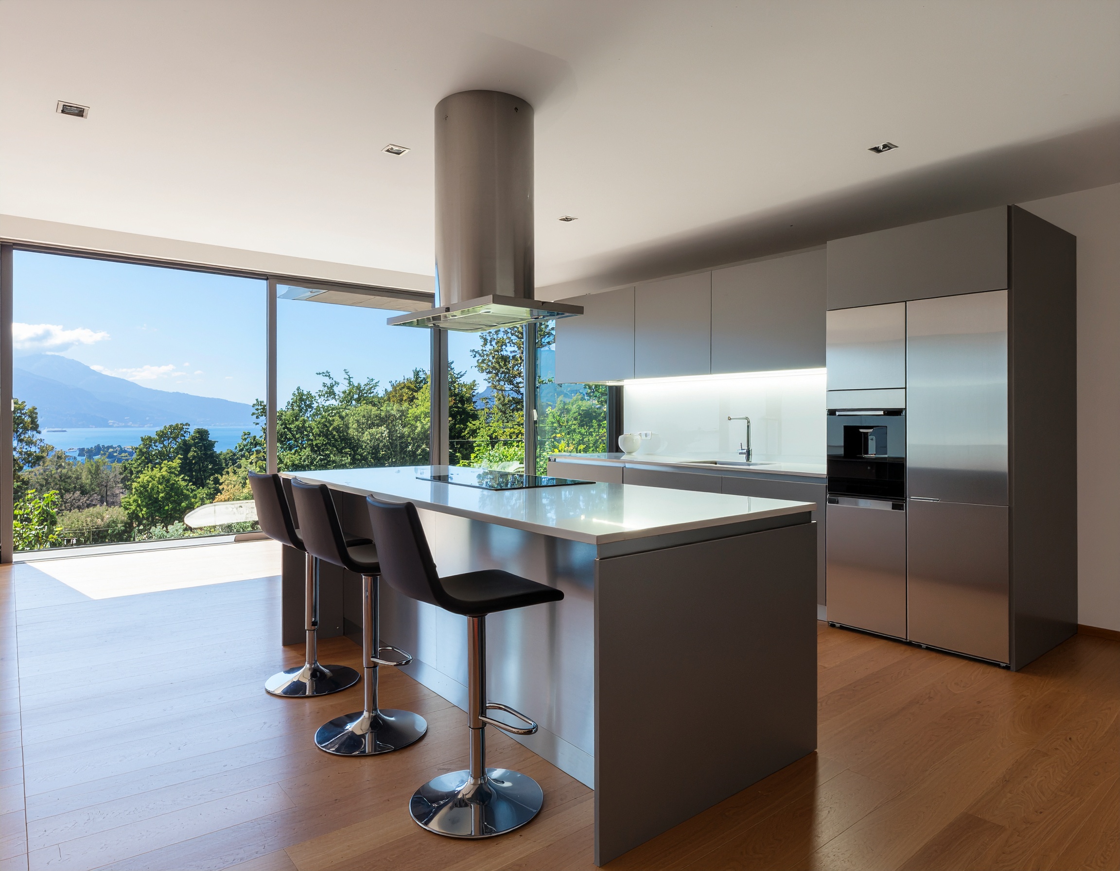 A high-end industrial kitchen featuring a stainless steel island with black leather barstools, a large cylindrical range hood, grey cabinetry, and floor-to-ceiling windows with a scenic lake and mountain view.