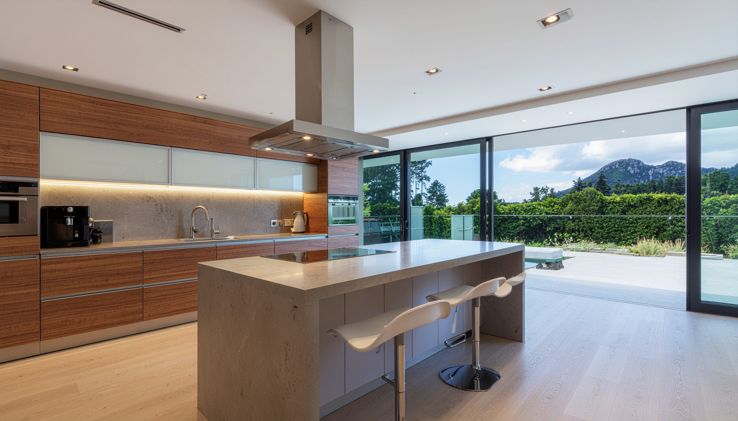 A spacious kitchen featuring warm wood cabinetry, a massive grey stone island with white barstools, and floor-to-ceiling glass doors opening to a lush garden and mountain landscape.