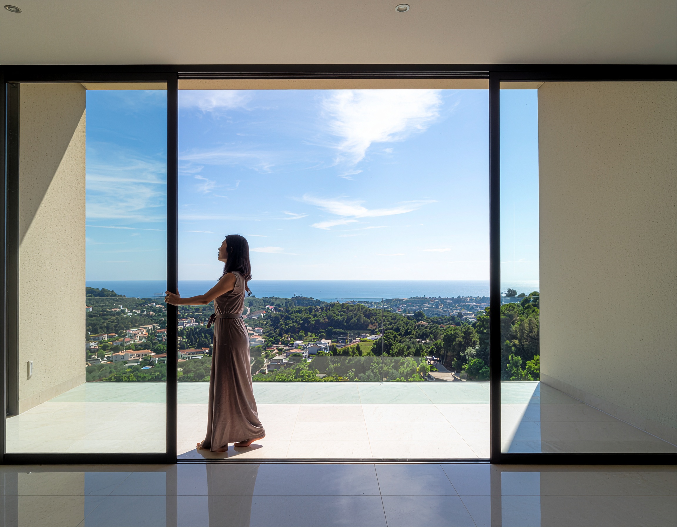 Full-body shot of a woman in a long flowing dress opening a large, black-framed sliding glass door, looking out at a sunny coastal landscape with a town in the valley below.