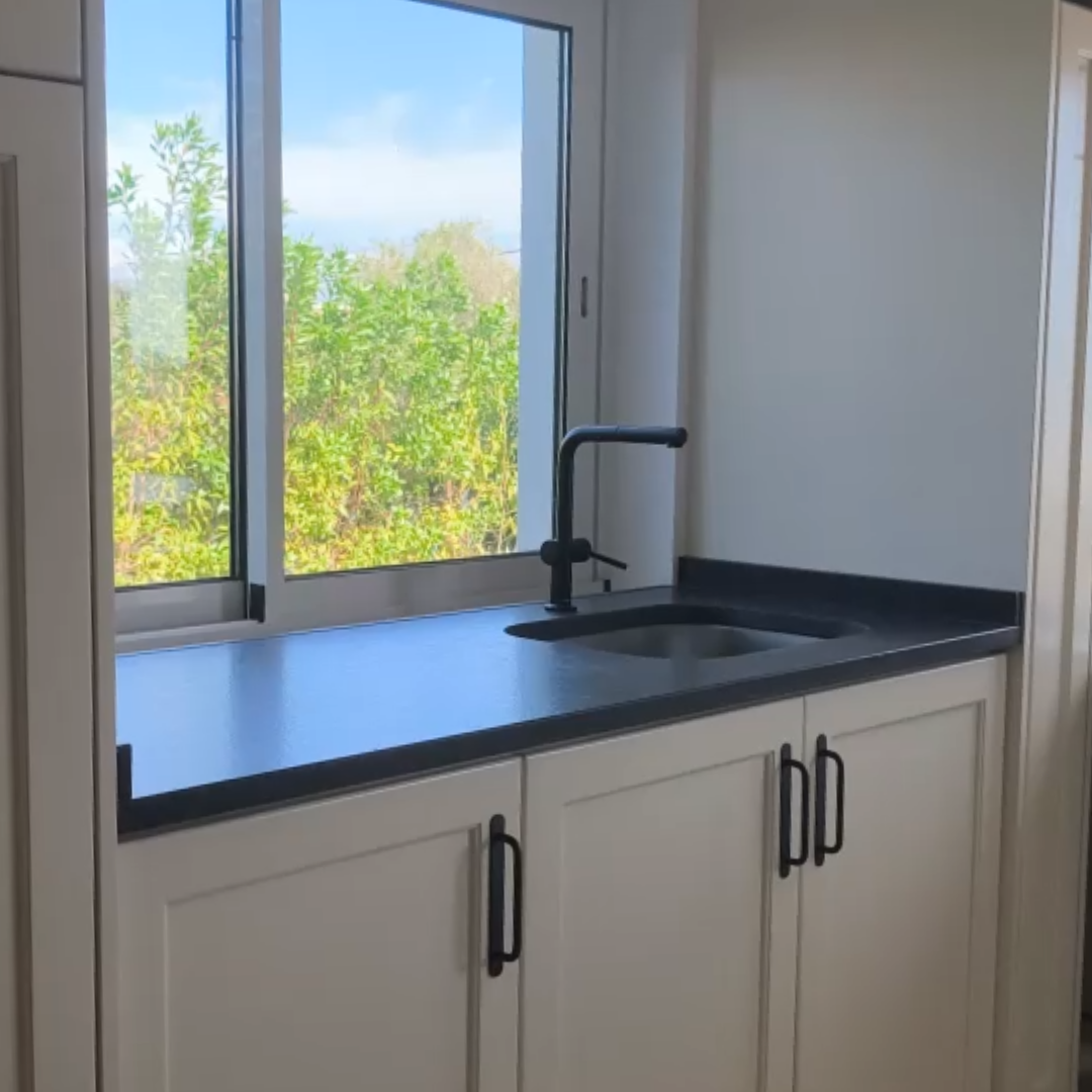 A close-up of a matte black kitchen countertop featuring an integrated sink and a matching black faucet. Below are white shaker-style cabinets with slim black handles. The sink is positioned in front of a bright window overlooking lush green trees.